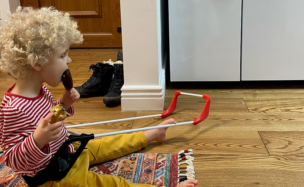 three year old girl sitting on the floor eating a chocolate ice cream pop. Wearing her belt cane her left two is hooked over the top of the cane frame.