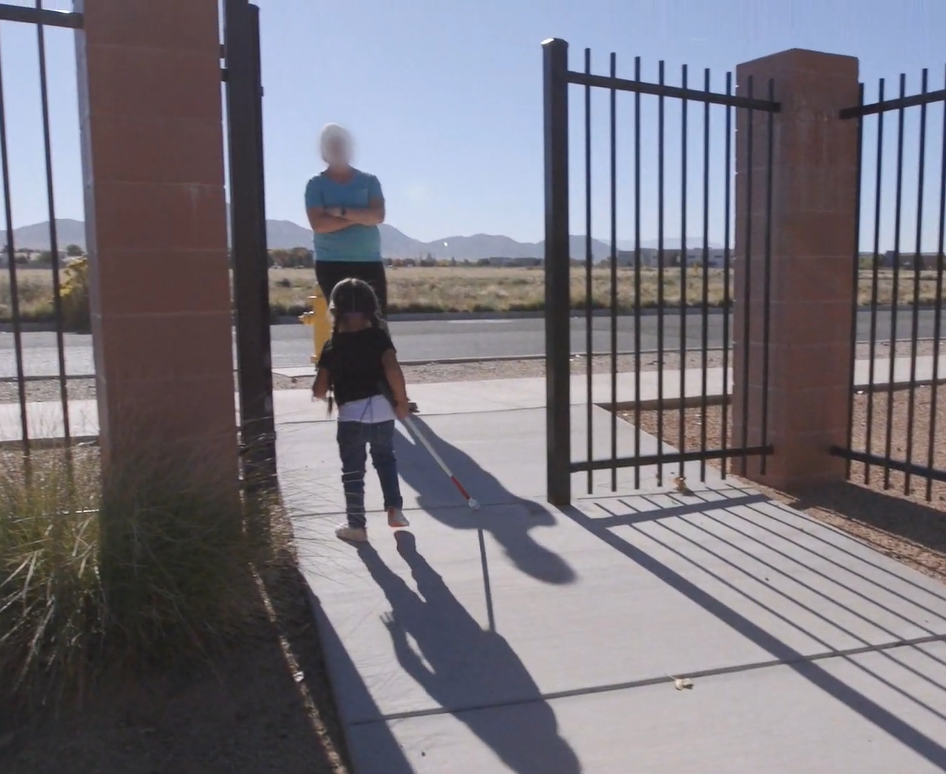 Child walking with white cane toward an adult by open black metal gate on sunny sidewalk, desert mountains in background.