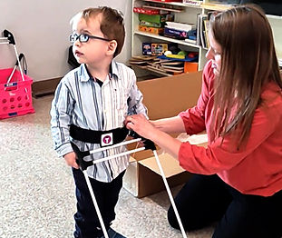 3-year-old boy wearing glasses stands as his teacher puts on his belt cane