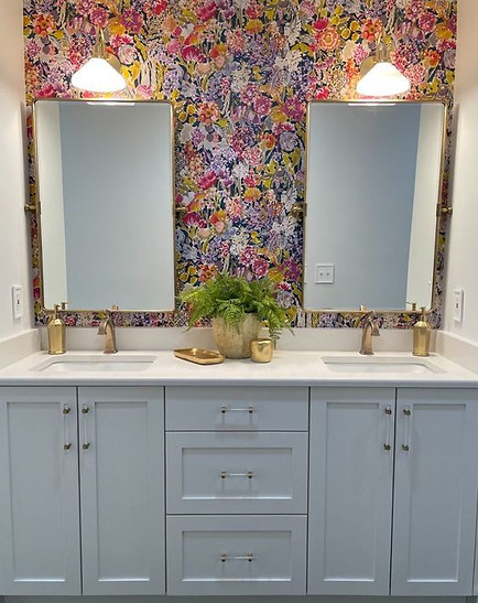 Vibrant floral wallpaper with rich blues, bright greens and pinks, and a light lavender is the background for this bathroom vanity. The vanity has two sinks with matching, symmetrical gold faucets and soap dispensers. Above them are gold-lined mirrors and sconces. In between the two sinks sits a fern plant, a gold water cup, and a gold catch-all dish.