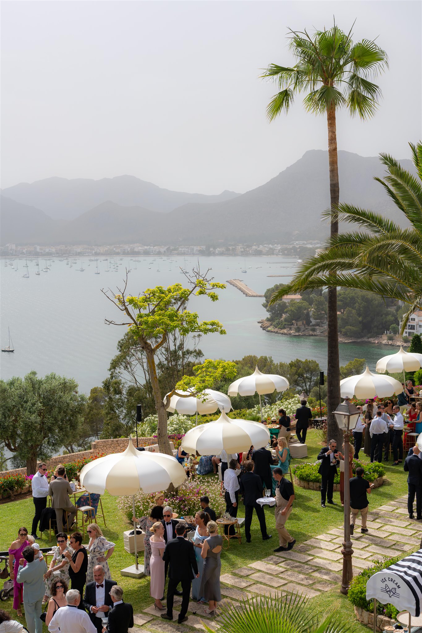 Outdoor wedding reception in La Fortaleza with round tables, white umbrellas and guests seated in a garden setting overlooking the bay of Pollenca