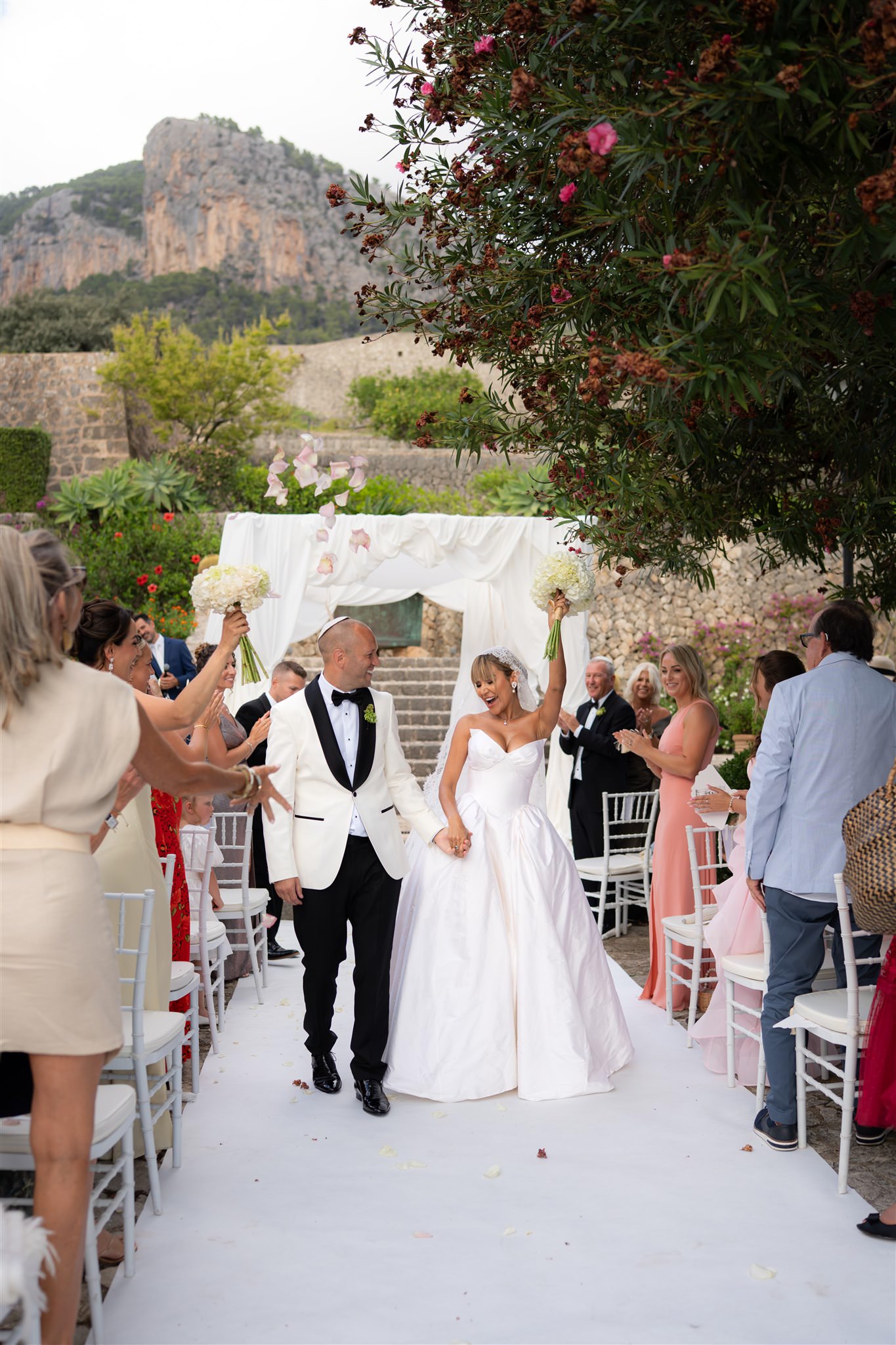 Bride and groom walking down a white aisle with guests standing on both sides with amazing mountains of Alaro in the background and a white chuppah