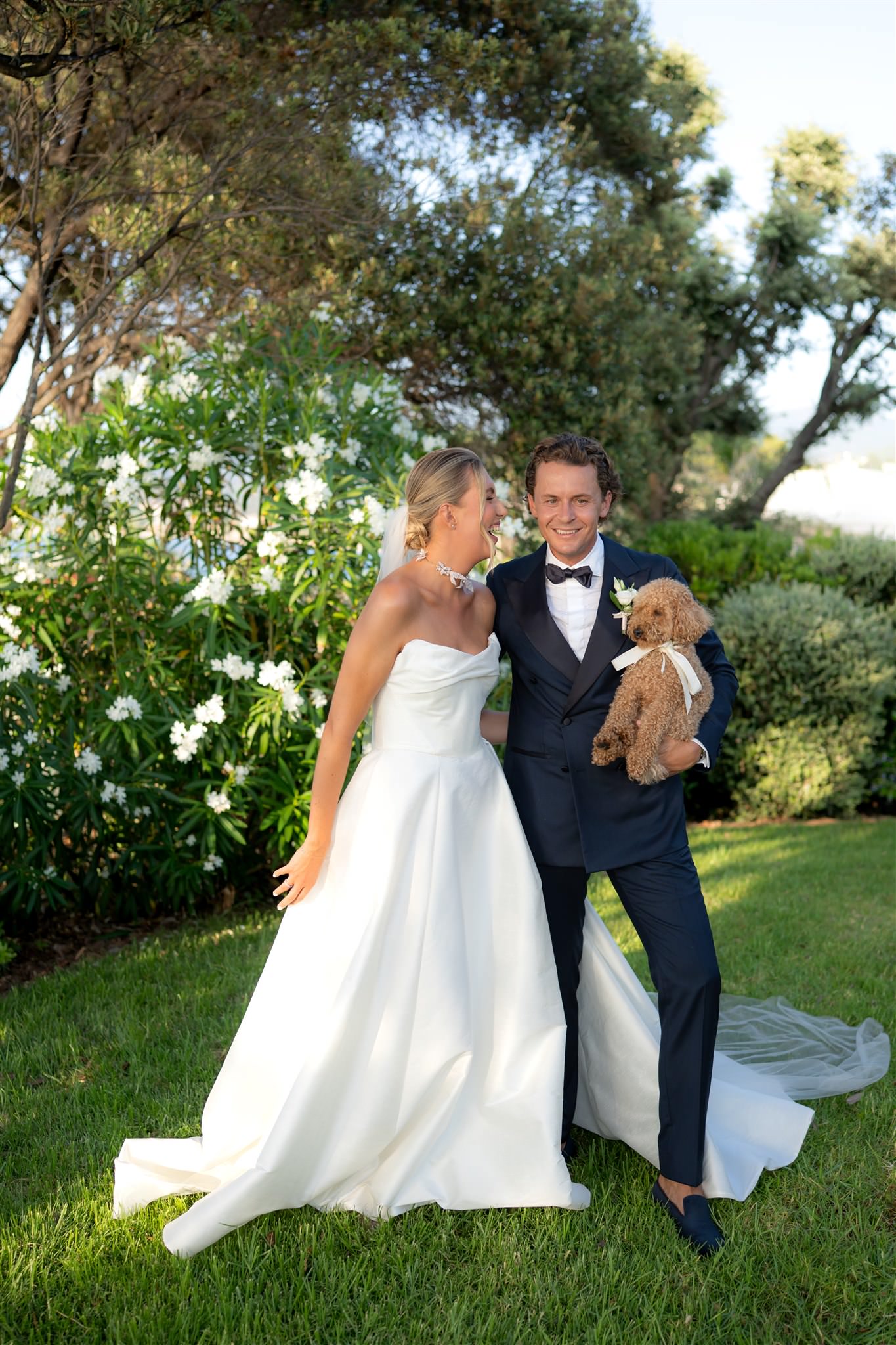Photograph of bride and groom standing together outdoors in a garden surrounded by greenery with their dog in Corsica