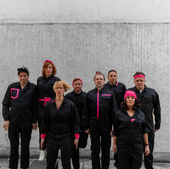 Gruppe in schwarzen Anzügen springt. Rosa Akzente, PRESSE, vor einer grauen Wand.