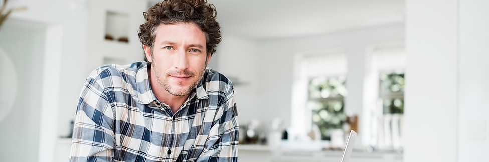 Homem na crise da meia-idade com cabelo cacheado, branco, sorriso tímido, apoiado no balcão olhando para frente.