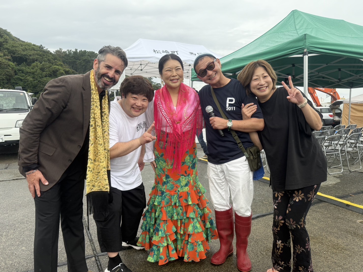 Cinco personas posan juntas junto a unas carpas de evento; una lleva un vestido de flamenco con estampado floral.