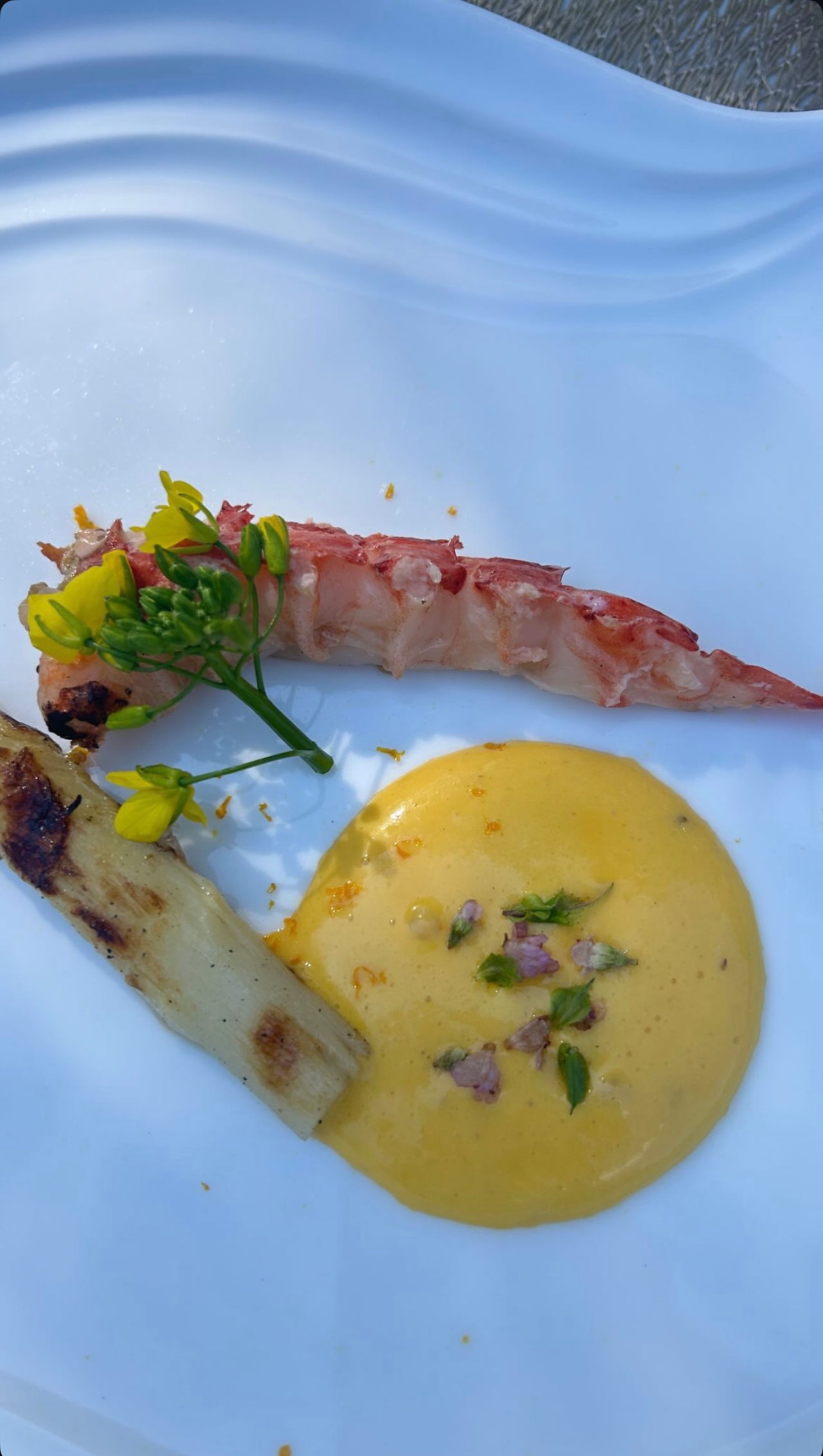 Un plato blanco con productos del mar, una verdura a la parrilla, salsa amarilla y flores pequeñas.
