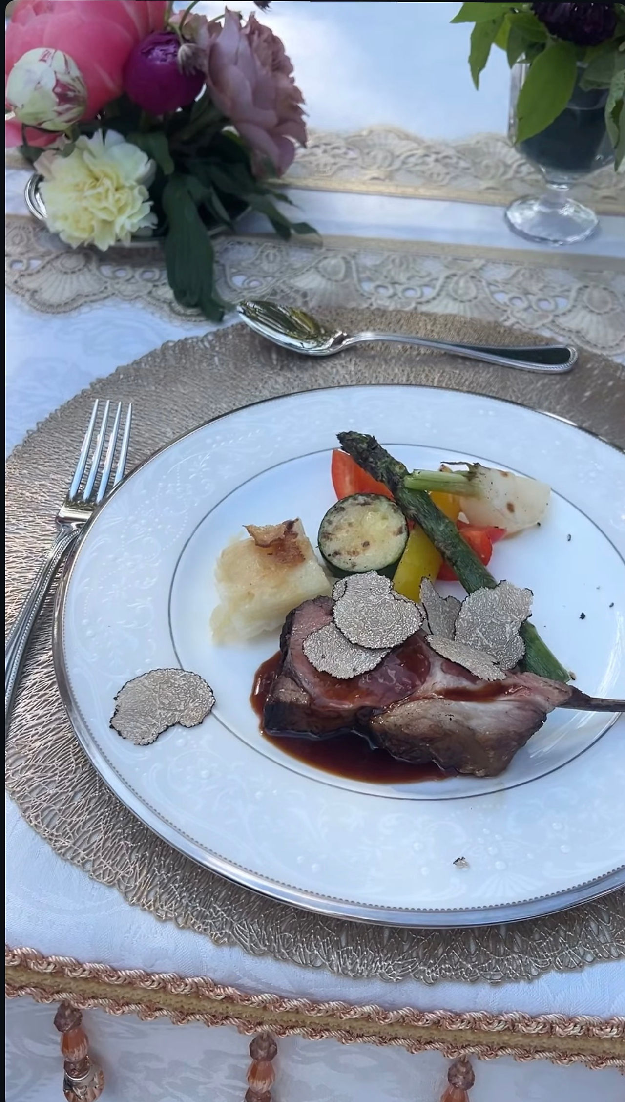 Un plato de comida con lonchas de carne, verduras y láminas finas de setas junto a unas flores.