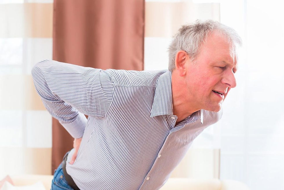 Elderly man clutching his lower back in pain, wearing a striped shirt, standing indoors with a brown curtain in the background.