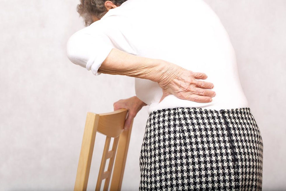 Elderly person in a white shirt and patterned skirt holding back in pain, leaning on a wooden chair in a neutral-toned room.