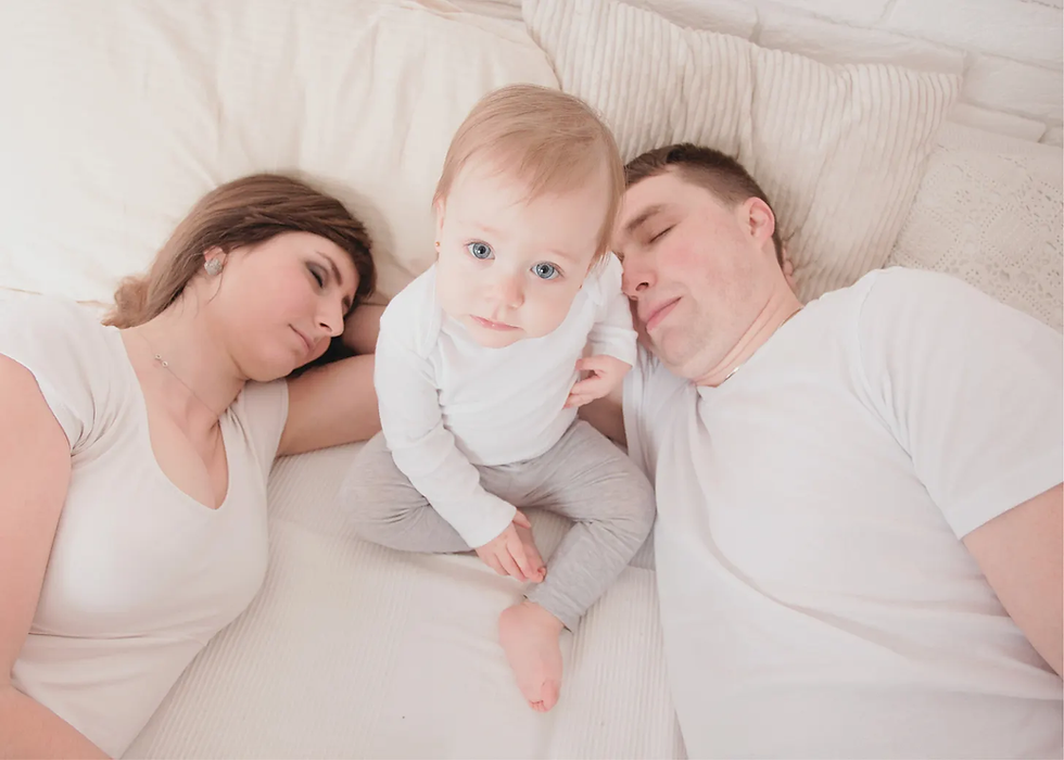 Woman and man are sleeping, a wide awake baby sits between them.