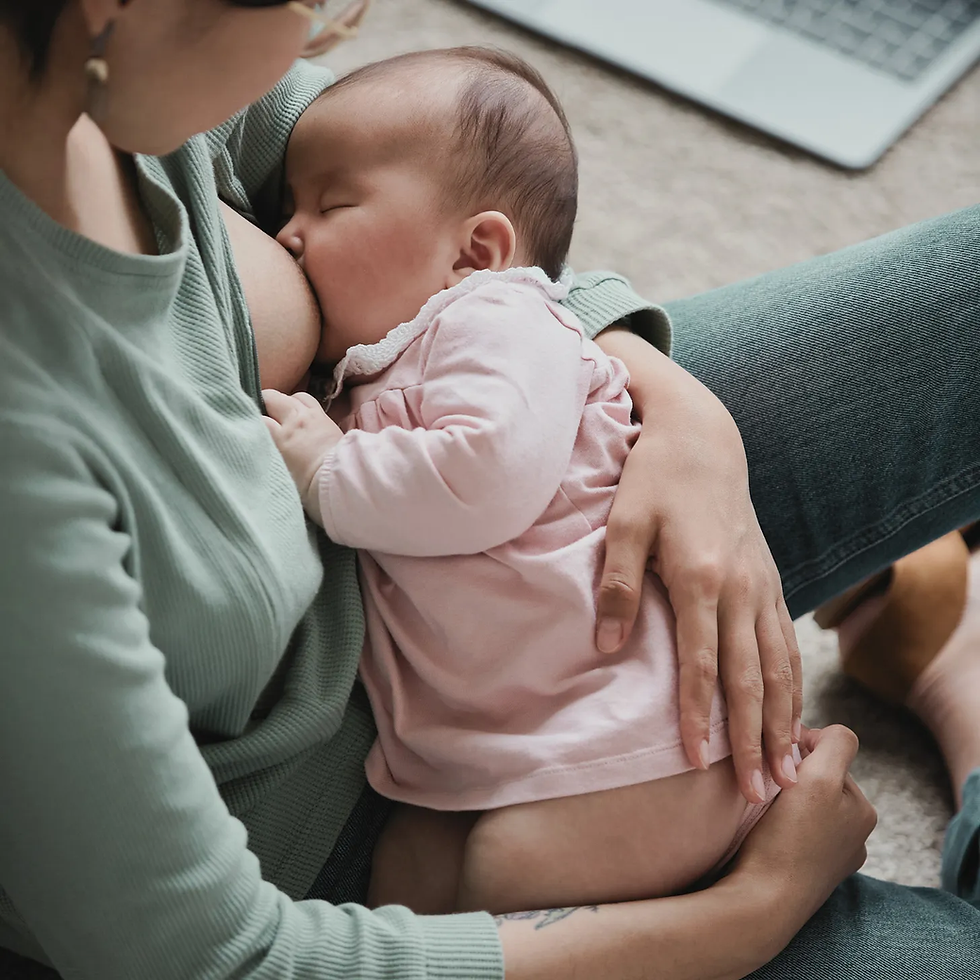 A woman sits on the floor nursing a baby. There is a laptop in the background.