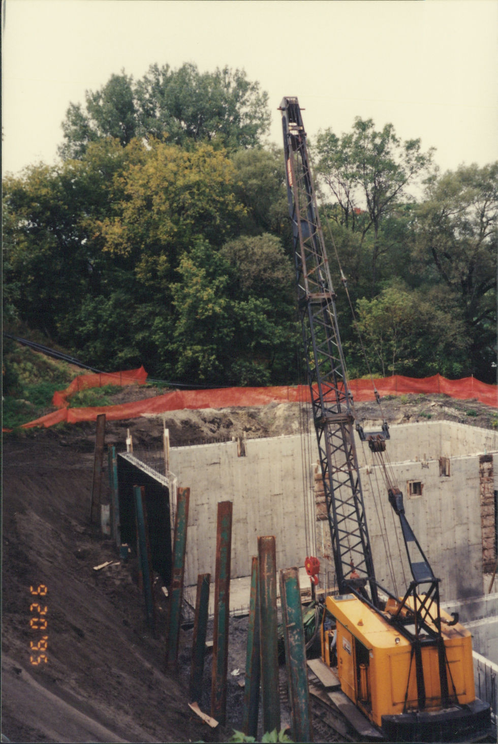 Grue de construction travaillant sur une structure en béton, arbres luxuriants derrière.