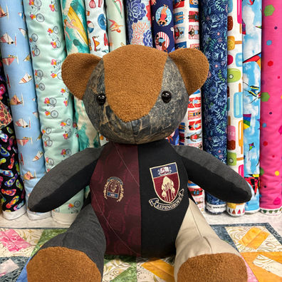 Patchwork teddy bear with a crest, sitting before fabric rolls.
