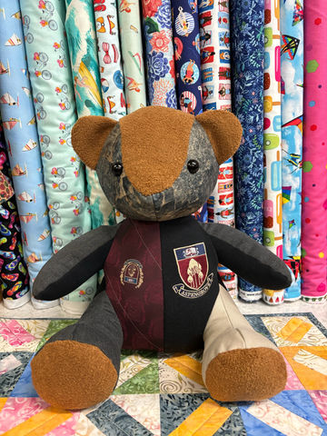 Patchwork teddy bear with a crest, sitting before fabric rolls.