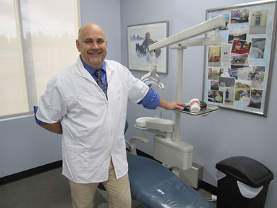 Smiling bald dentist stands beside dental chair in bright office.
