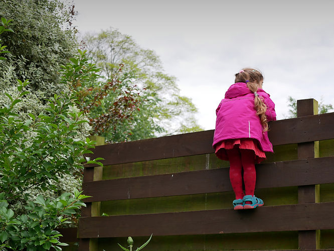 girl with a pink jacket and red tights looking out from a wooden fence, plants at the side
