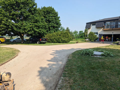 Dirt driveway curves toward a house, cars and trees, bright summer day. Tri City Paving Inc