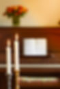 Two thin candles in candle stick holders burning against blurry piano and bouquet of flowe
