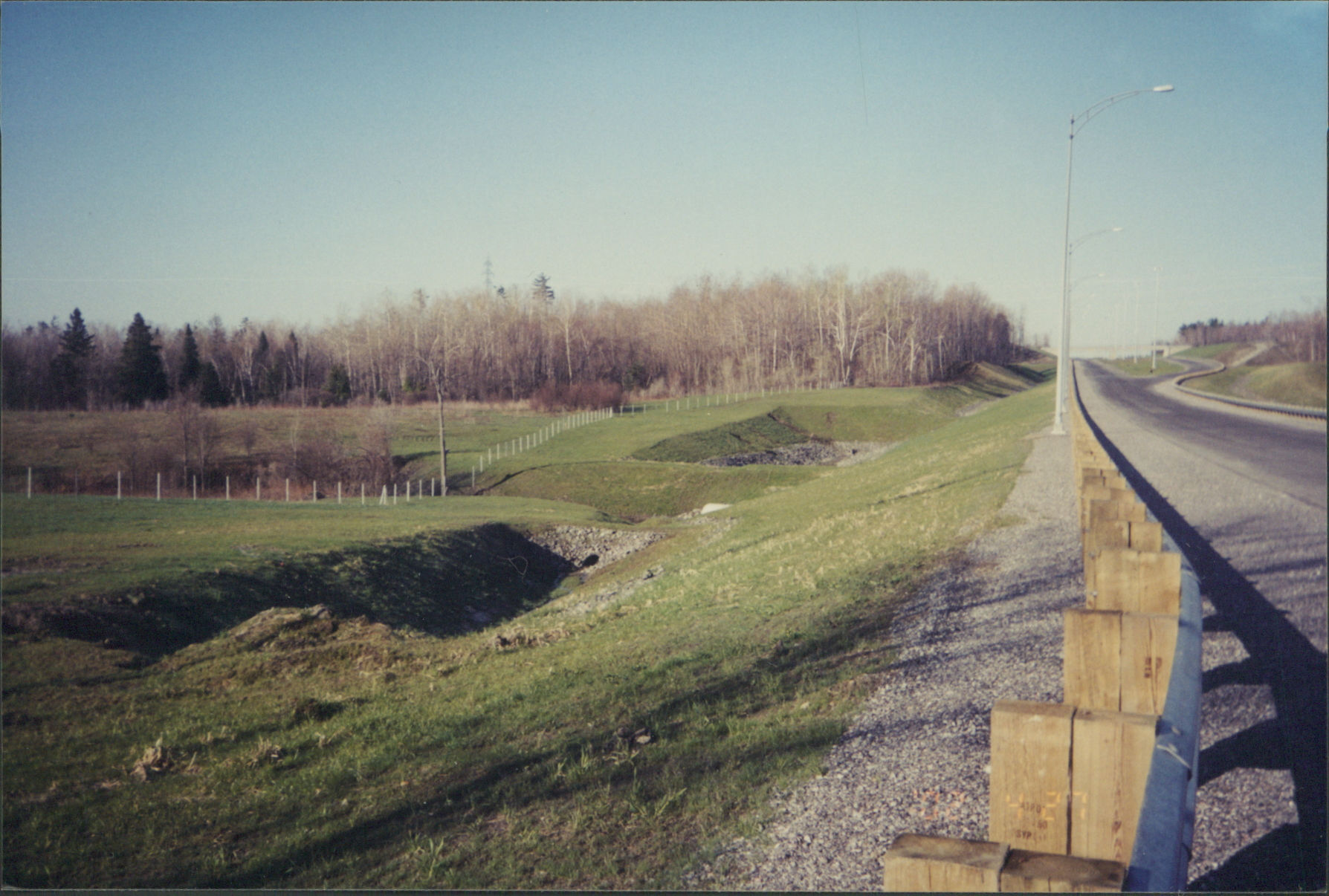Route avec glissière de sécurité, talus enherbé et arbres lointains.