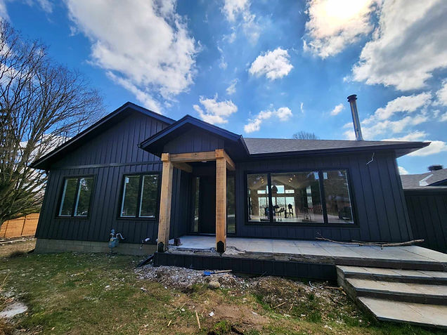 Modern dark-sided house with wooden porch pillars and large windows under a blue sky.