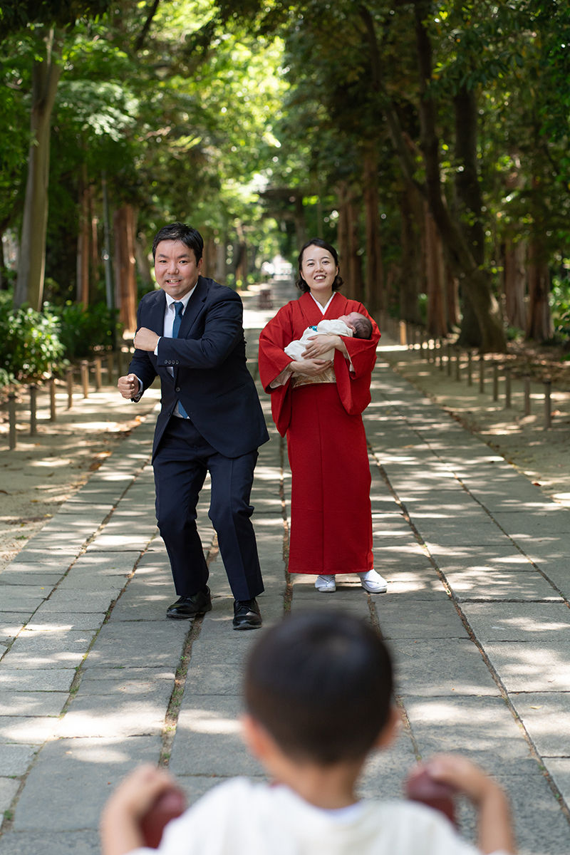 越谷市・久伊豆神社でのお宮参りの出張撮影。木立の参道で、紺のスーツの父と赤い着物の母が赤ちゃんを抱き、手前の子どもに向かって笑顔で立つ家族写真。