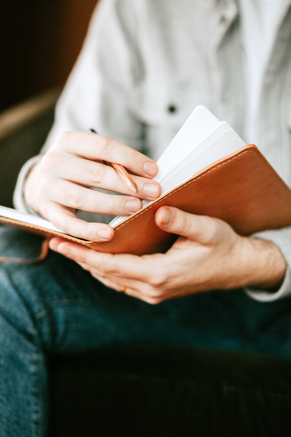 Hands holding an open brown notebook, turning pages with a pencil. The setting is casual with a cozy, soft-focus background.