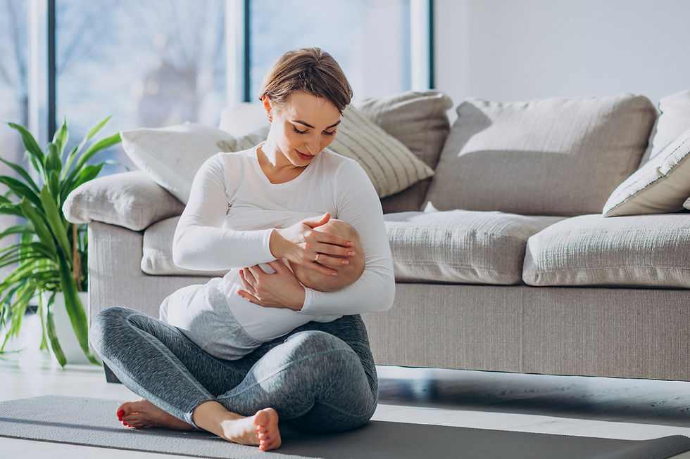Mutter stillt Baby auf einer Yogamatte vor einem grauen Sofa. Helle, sonnige Wohnzimmeratmosphäre, grüne Pflanze im Hintergrund.