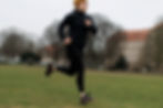 Person joggt auf Wiese, trägt schwarze Kleidung und gelbe Mütze. Hintergrund Bäume und Gebäude mit rotem Dach. Bewölkter Himmel.