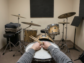 A quiet private drum practice room with an acoustic drum kit set up. Only the drummer’s hands and lower arms are visible, resting on the snare while holding still drumsticks. The room is neutral and uncluttered, with no motion, no performance, and no implied action.