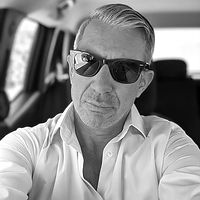 A confident man with short, neatly combed hair wearing sunglasses and a white shirt, seated in a car, providing a testimonial for DrumOrama.blog.
