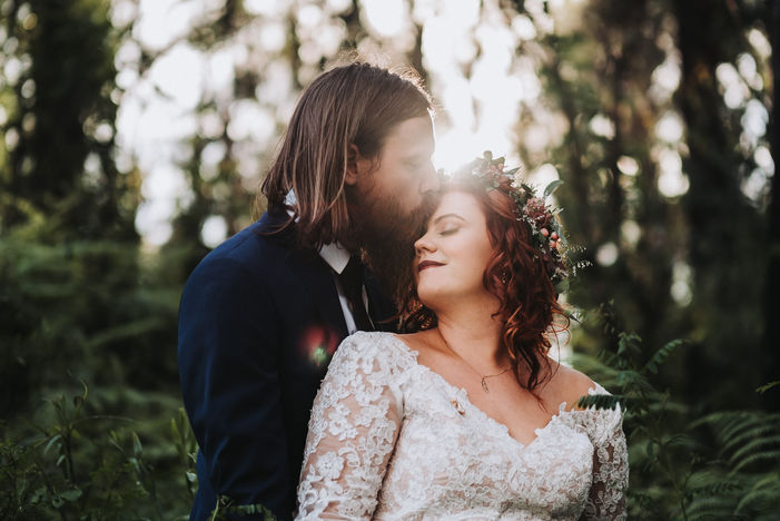 Groom kissing bride on forehead surrounded by forest and filtered sunlight, captured by wedding photographer Oscar Colman