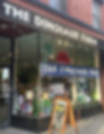 Storefront of "The Dinosaur Farm" with toys and books visible through large windows. A-frame sign outside. Black and red decor.