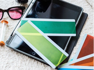 Black headphones resting on a tablet and stylus on a cream rug, surrounded by colourful swatch cards, pink-tinted glasses, and a small hourglass