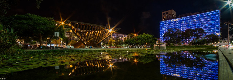 Wide-angle night panorama of Rabin Square, showing the illuminated Tel Aviv City Hall building glowing in blue and the Holocaust Memorial under the stars.