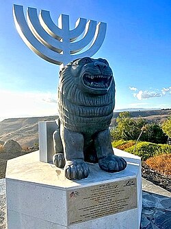 A modern bronze lion sculpture standing proudly beneath a metal menorah symbol at the entrance plaza of the Ein Keshatot heritage site.