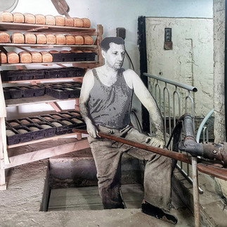A museum display featuring a cutout figure of a young pioneer worker unloading freshly baked loaves of bread from industrial kitchen racks.