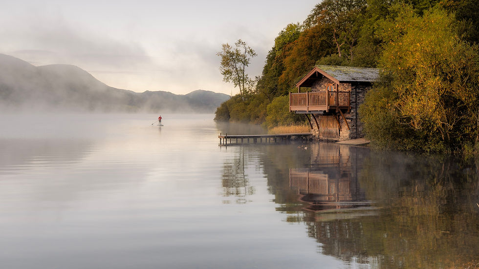 Wide-angle view of a misty Lake District lake at sunrise with a lone paddle boarder near a boathouse.