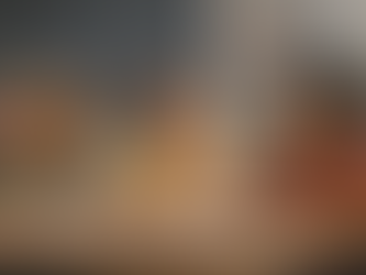 Three primary-aged children sitting at a table in a classroom, writing in exercise books together, with a chalkboard and bookshelves in the background.