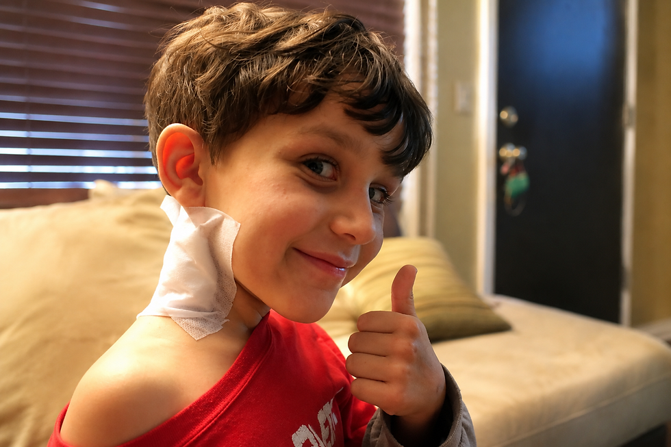A boy with a sweet smile and a large bandage on his neck giving a thumbs up