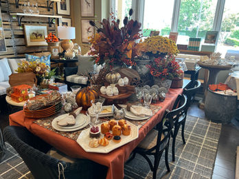Table set with a fall floral centerpiece, pumpkins, and place settings near a window.