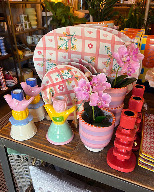 A display of plaid dishes in pastel colors, small potted pink orchids in striped planters, and whimsical candle sticks with colorful birds and flowers.