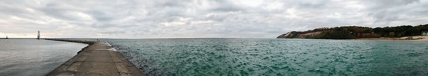 A panoramic lake view with a breakwater path leading out to a lighthouse. The water on the left is calm and the water on the right is rough. 