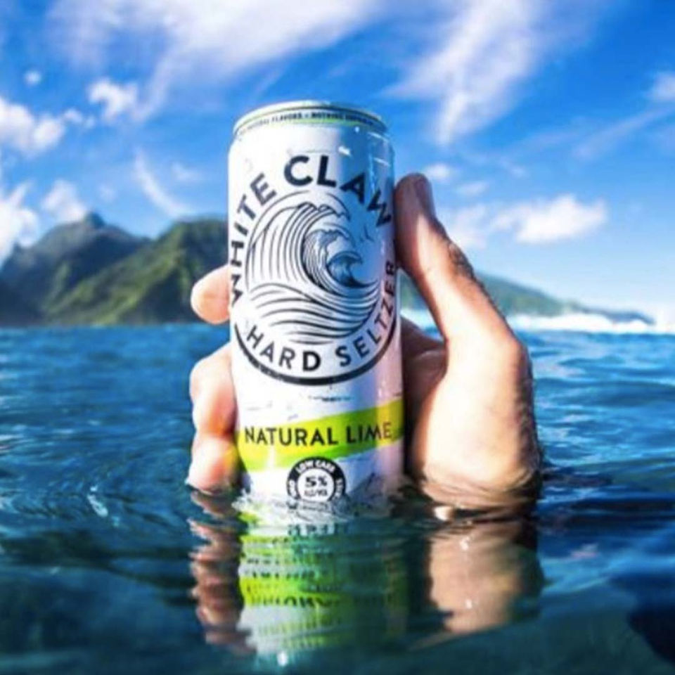 A hand lifts a White Claw Natural Lime hard seltzer from ocean water, captured in a bright, beachside setting for a summer lifestyle campaign.