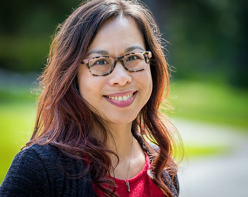 Headshot of Asian American woman with long wavy hair and glasses, wearing a red top and dark gray cardigan against a green and white background. 