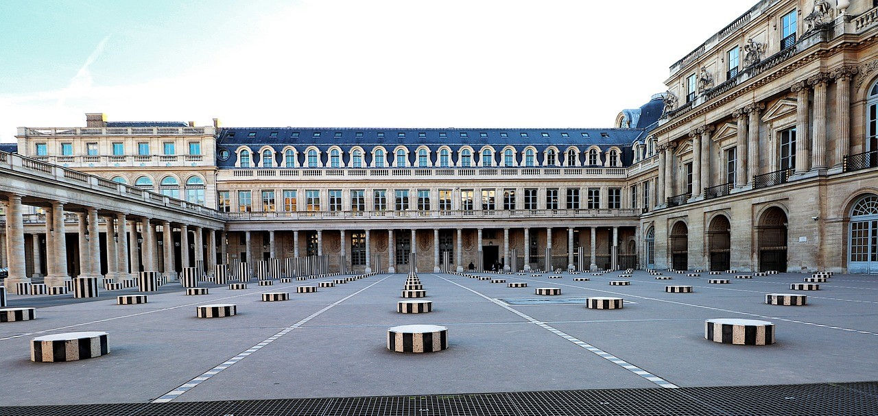 A courtyard with a series of black and white striped columns of varying heights is set against a grand, multi-story building with arched windows and a slate roof.