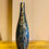 Thumbnail: Tall, slender decorative vase with a shimmering mosaic tile finish in shades of blue, gold, and bronze, sitting on a polished