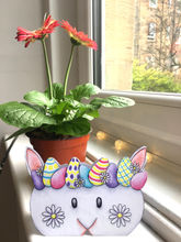 A hand-drawn, coloured-pencil illustration, Easter card. The card is in the shape of a white rabbit or Easter Bunny's head, wearing a crown of pastel-coloured, patterned Easter eggs on it's head. It has a daisy flower on each cheek. It is displayed stood up, facing forward, with some potted red flowers behind it in the background.