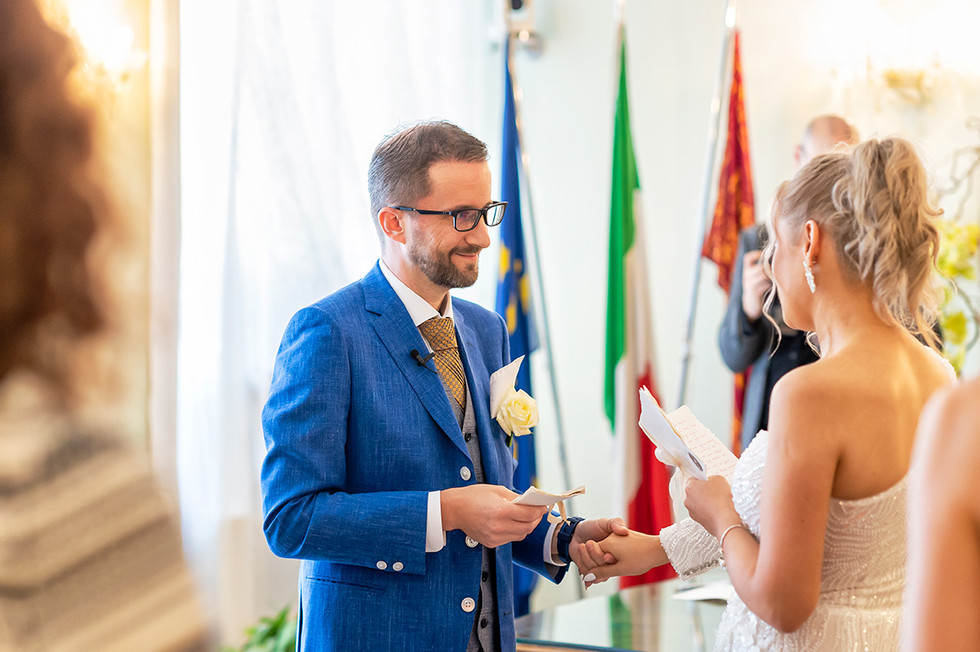 Close-up of the couple's hands clasped together, exchanging rings, with the elegant interior of Palazzo Cavalli as a backdrop.