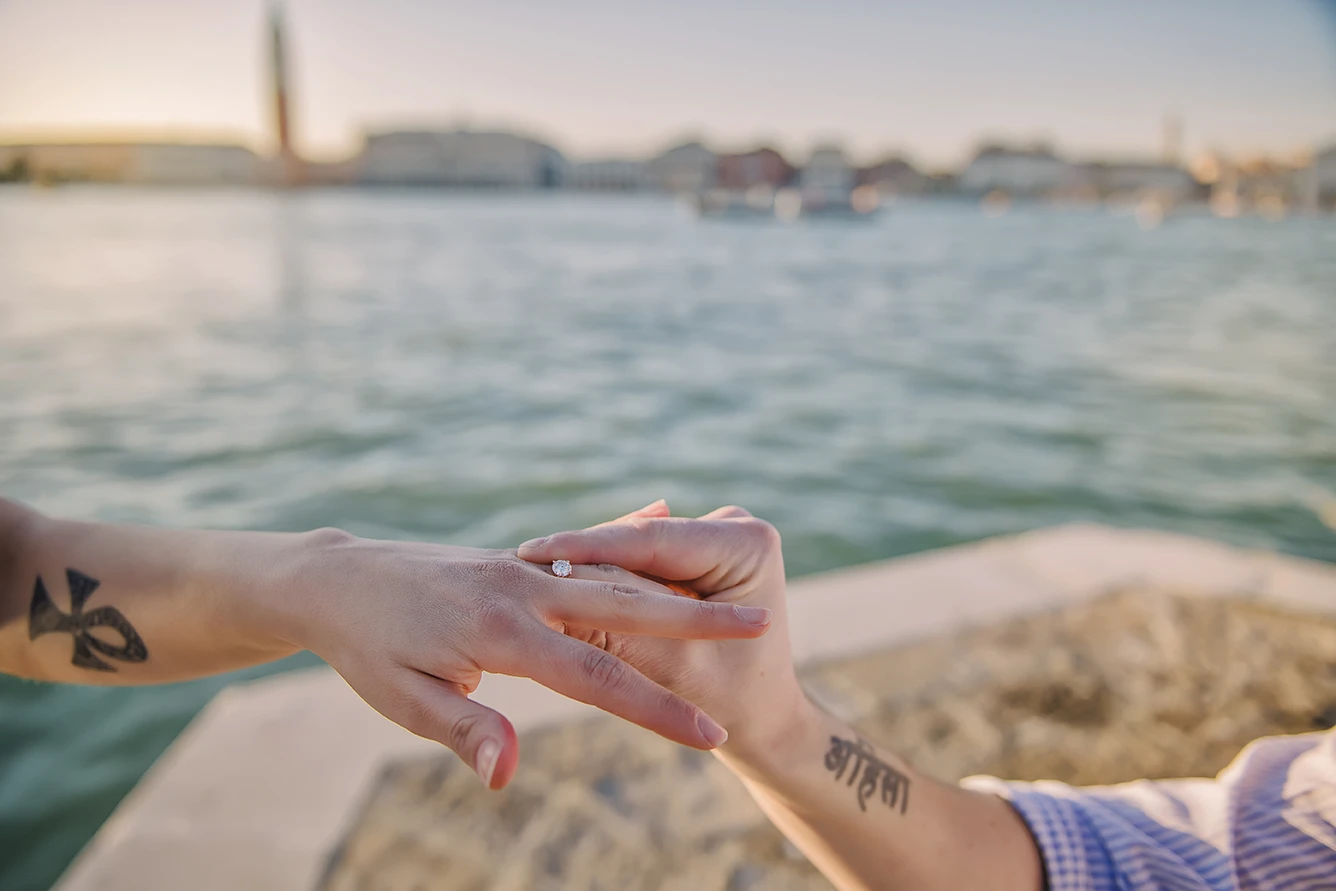Close-up of Maria's hands as she lovingly touches Barth's shoulder while looking down at the ring with teary eyes of happiness against the backdrop of the Venice lagoon.